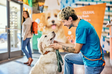 Dogtopia employee in Dogtopia location lobby petting white dog with dog's female owner in background leaving the facility